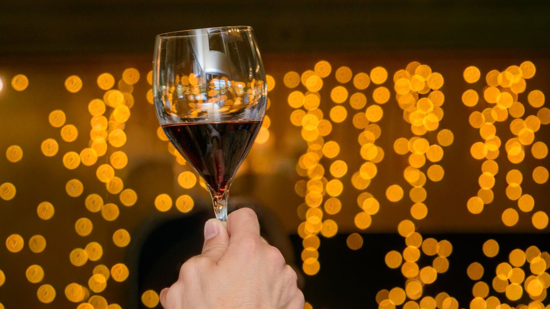 A hand holding a red wine glass against warm bokeh lighting.
